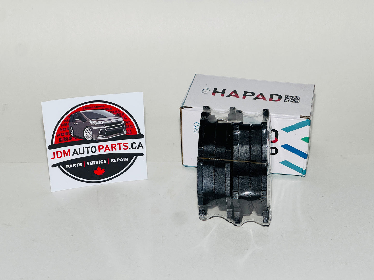 A black automotive brake pad with a clear outer cover, displayed next to its white HAPAD box and a JDM Auto Parts business card on a light gray background.