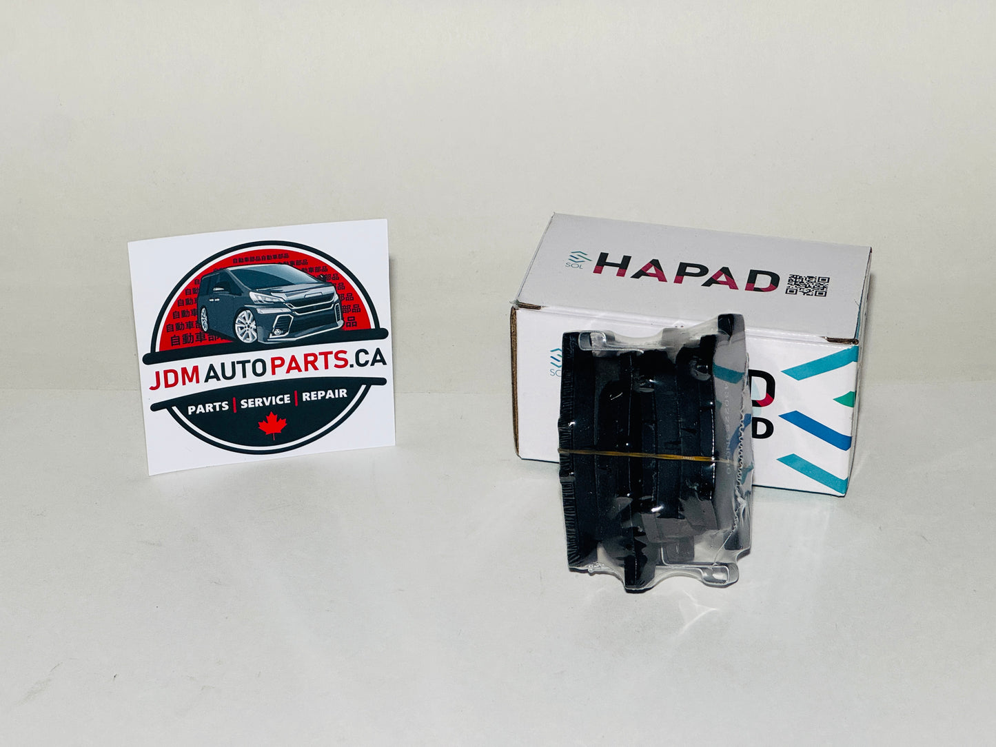 A black automotive part in clear plastic packaging stands in front of a white HAPAD box on a light gray background, with a JDM Auto Parts card on the left.