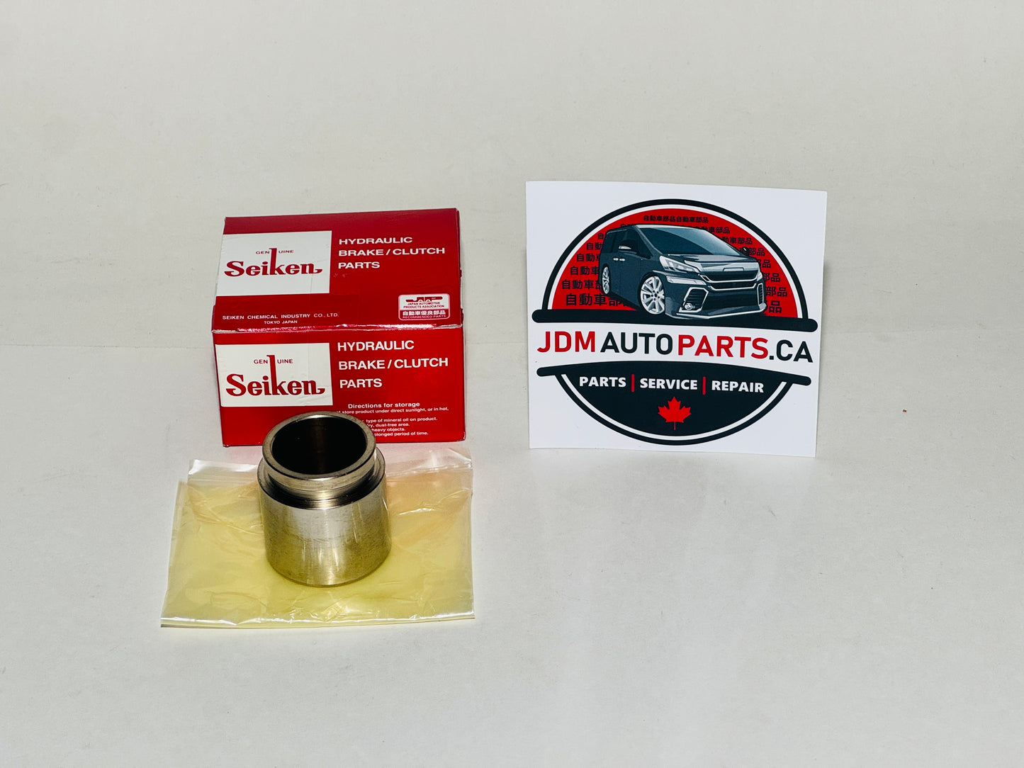 A Seiken hydraulic brake piston parts component in a clear plastic bag with a metal cylinder, shown next to its red Seiken retail box and a JDM Auto Parts logo sticker on a white background.