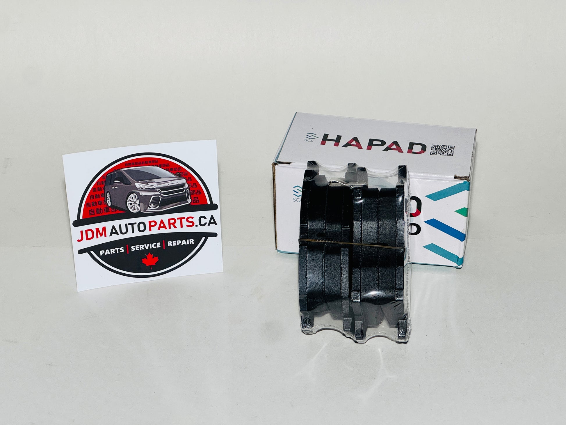 A black automotive brake pad with a clear outer cover, displayed next to its white HAPAD box and a JDM Auto Parts business card on a light gray background.