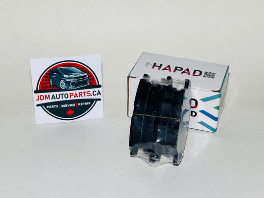 A black automotive brake pad set in a clear plastic case, shown next to its white HAPAD box and a JDM Auto Parts business card on a white background.