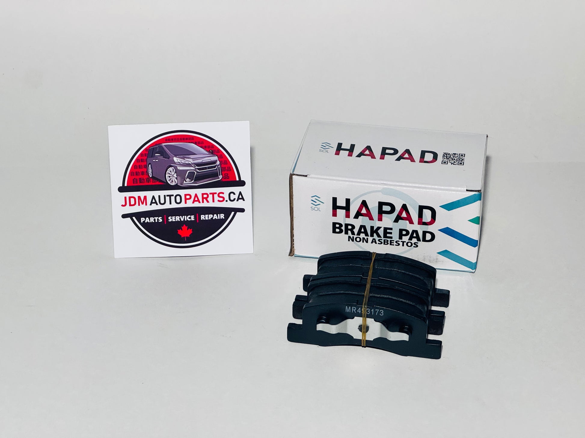 A pair of black HAPAD brake pads next to their white and blue retail box, with a JDM Auto Parts Canada sticker on a plain light gray background.