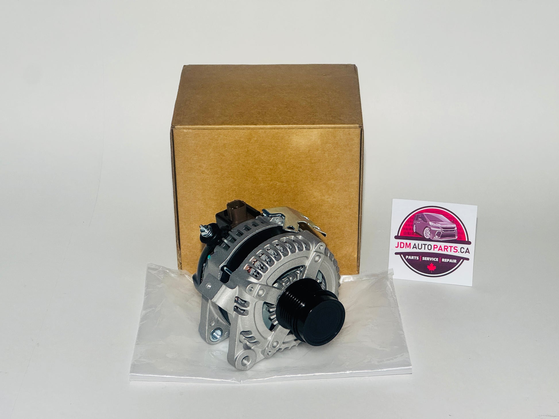 Car alternator on a white surface with a cardboard box and JDM Auto Parts CA label in the background. 2008-2014 TOYOTA ALPHARD AND VELLFIRE ALTERNATOR FOR 2.4L 2AZ-FE MODELS ANH20 ANH25 