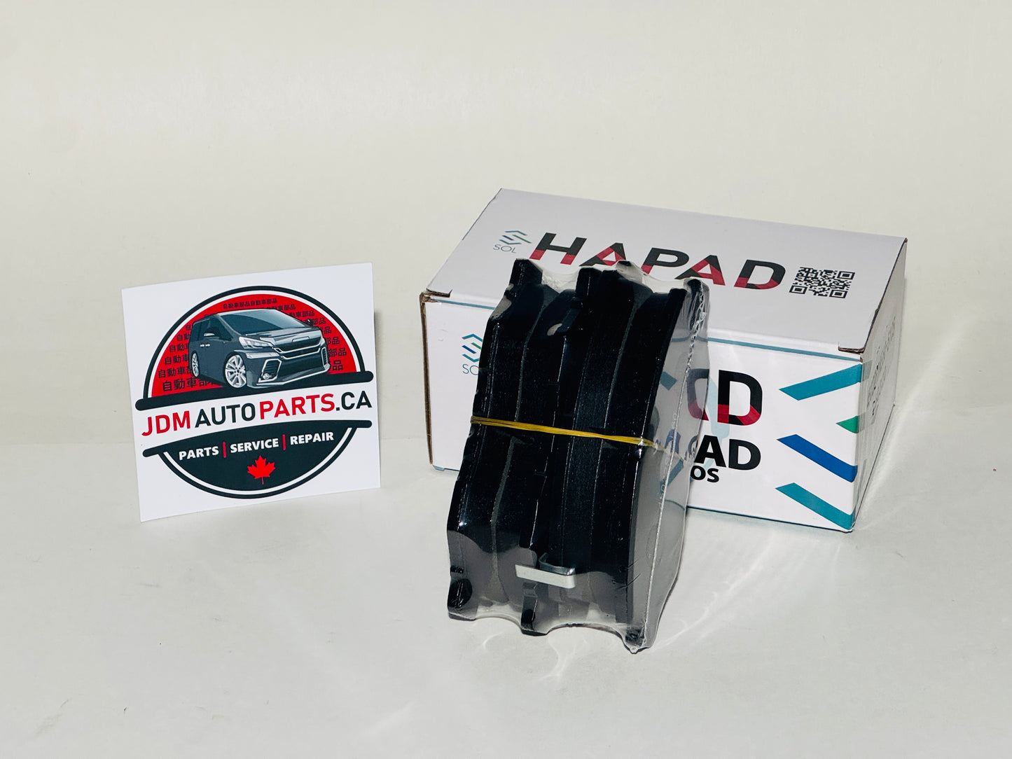 A black automotive part in a clear plastic sleeve stands in front of its white HAPAD box, with a JDM Auto Parts card on a plain white background.
