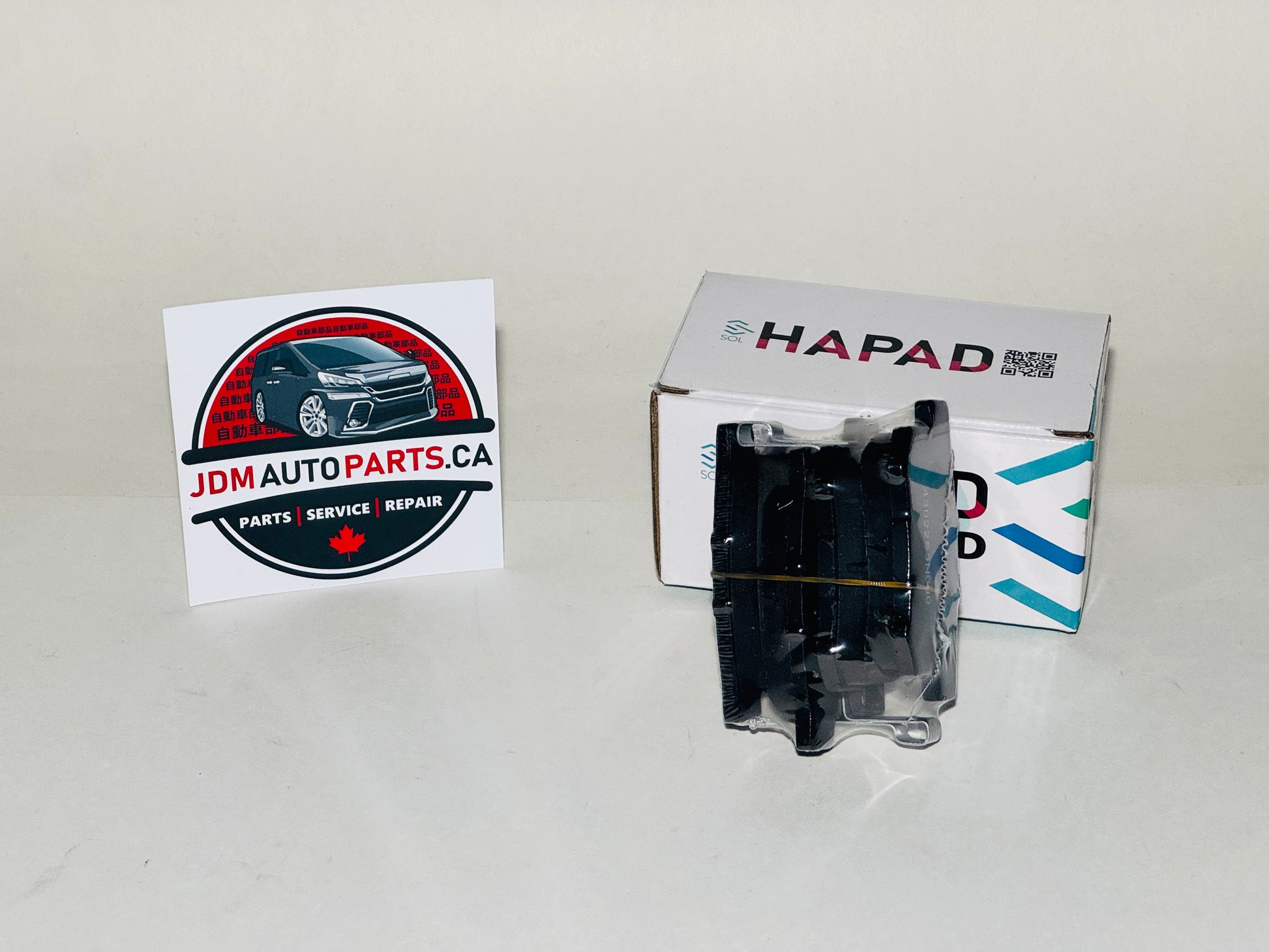 A black automotive part in clear plastic packaging stands in front of a white HAPAD box on a light gray background, with a JDM Auto Parts card on the left.